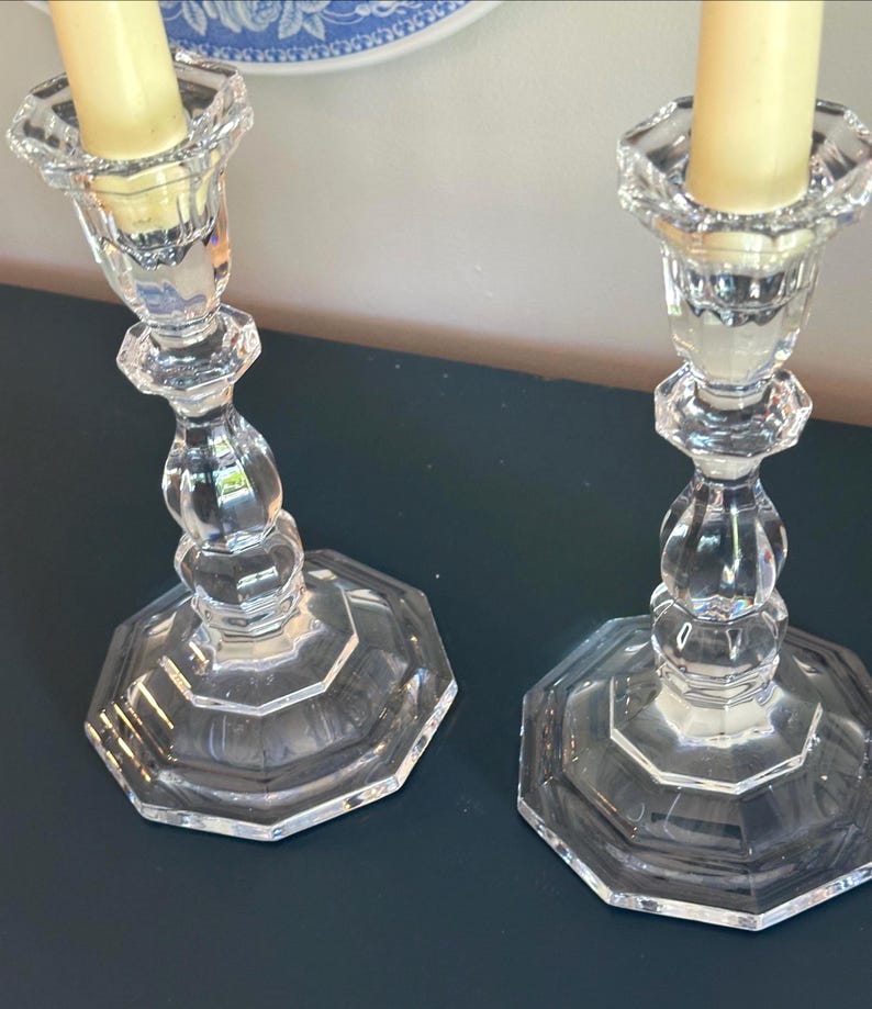 May include: A pair of clear glass candle holders with an octagonal base and a faceted, tiered stem design. Each holder features a scalloped top rim and contains a single, smooth, cream-coloured taper candle inserted into the central socket.