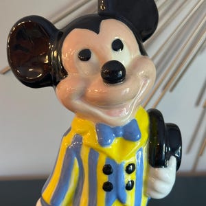 May include: A ceramic figurine of Mickey Mouse wearing a yellow and blue striped jacket with a blue bow tie and holding a black hat.