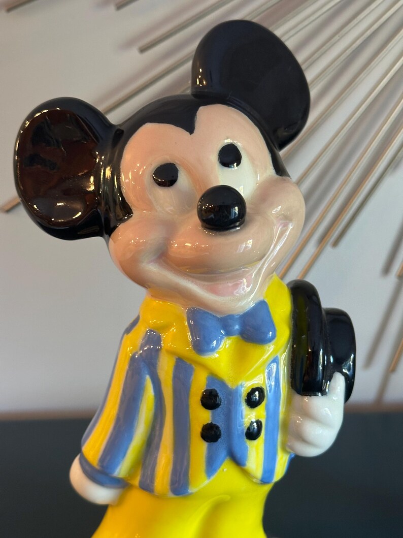 May include: A ceramic figurine of Mickey Mouse wearing a yellow and blue striped jacket with a blue bow tie and holding a black hat.