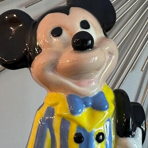 May include: A ceramic figurine of Mickey Mouse wearing a yellow and blue striped jacket with a blue bow tie and a black hat.