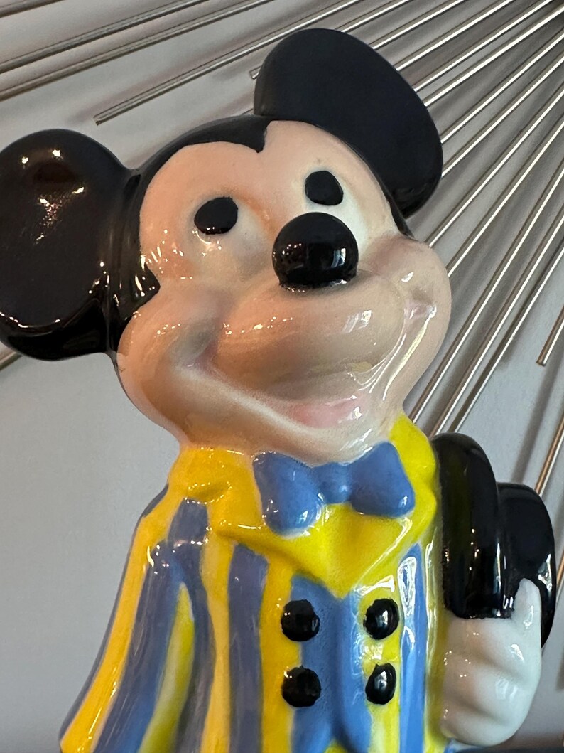 May include: A ceramic figurine of Mickey Mouse wearing a yellow and blue striped jacket with a blue bow tie and a black hat.