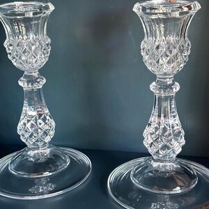 May include: A pair of clear glass candle holders featuring a faceted diamond-cut pattern on the upper cup and the central stem. Each holder stands on a wide, smooth, circular base, with a flared top rim designed to securely hold a standard taper candle.