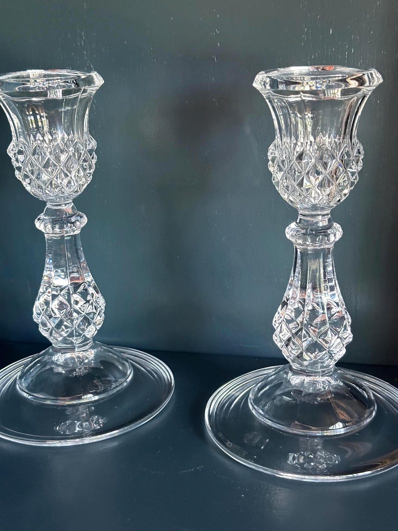 May include: A pair of clear glass candle holders featuring a faceted diamond-cut pattern on the upper cup and the central stem. Each holder stands on a wide, smooth, circular base, with a flared top rim designed to securely hold a standard taper candle.