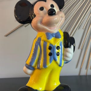 May include: A ceramic figurine of Mickey Mouse wearing a yellow and blue striped suit with a blue bow tie and holding a black hat.