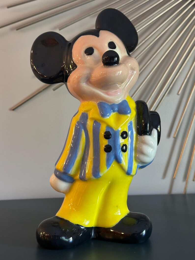 May include: A ceramic figurine of Mickey Mouse wearing a yellow and blue striped suit with a blue bow tie and holding a black hat.