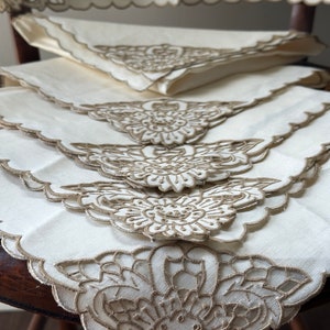 May include: Set of four white linen napkins with delicate brown embroidered floral designs. The napkins are folded and stacked on a wooden chair.