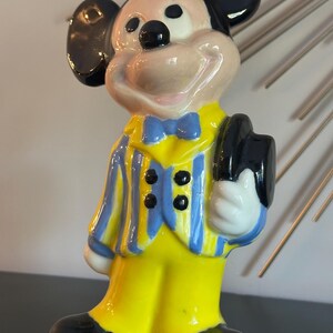 May include: A ceramic figurine of Mickey Mouse wearing a yellow suit with blue and yellow stripes, a blue bow tie, and a black hat. He is smiling and holding his hat in his right hand.