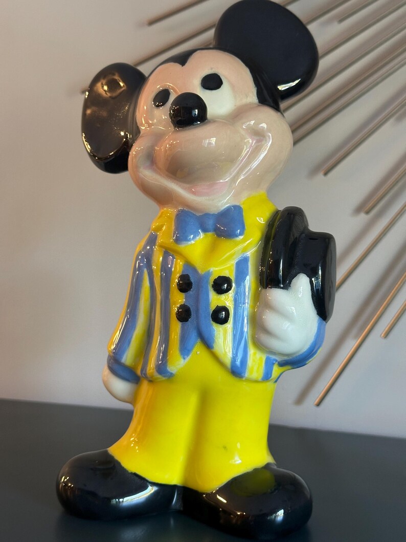 May include: A ceramic figurine of Mickey Mouse wearing a yellow suit with blue and yellow stripes, a blue bow tie, and a black hat. He is smiling and holding his hat in his right hand.