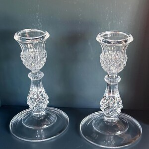 May include: A pair of clear glass candle holders featuring a faceted diamond-cut pattern on the upper cup and the lower stem. Each holder has a wide, smooth circular base and a flared top rim designed to securely hold a standard taper candle.