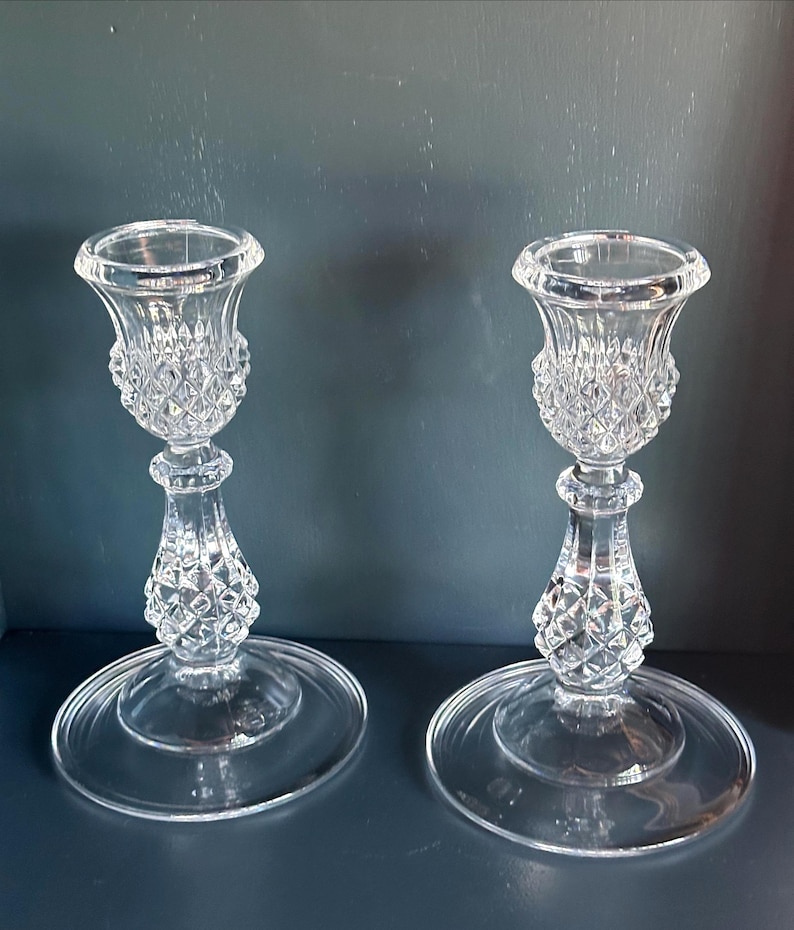 May include: A pair of clear glass candle holders featuring a faceted diamond-cut pattern on the upper cup and the lower stem. Each holder has a wide, smooth circular base and a flared top rim designed to securely hold a standard taper candle.