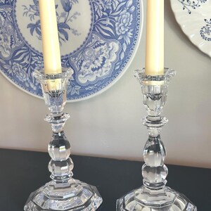May include: A pair of clear glass candle holders with a faceted, multi-sided base and a central stem featuring rounded, bulbous sections. Each holder supports a tall, slender, cream-coloured taper candle, showcasing a reflective, transparent finish.