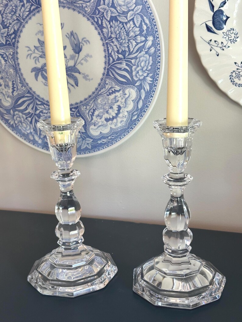 May include: A pair of clear glass candle holders with a faceted, multi-sided base and a central stem featuring rounded, bulbous sections. Each holder supports a tall, slender, cream-coloured taper candle, showcasing a reflective, transparent finish.