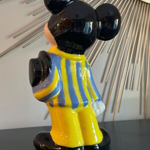 May include: A ceramic figurine of Mickey Mouse wearing a blue and yellow striped shirt and black shoes. He is standing with his back to the camera and his right hand is raised.