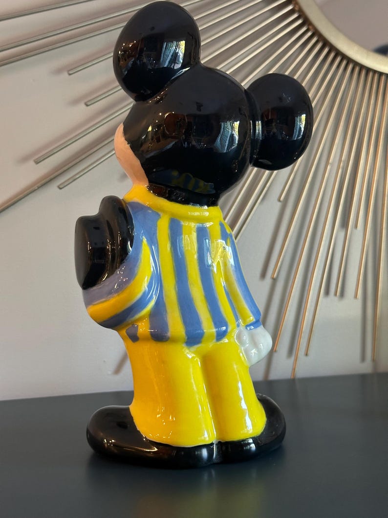 May include: A ceramic figurine of Mickey Mouse wearing a blue and yellow striped shirt and black shoes. He is standing with his back to the camera and his right hand is raised.