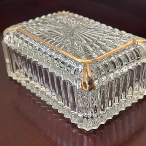 May include: A rectangular clear glass trinket box featuring a faceted, ribbed texture on the sides and lid. The lid is accented with a thin, metallic gold-colored trim along its edges, highlighting the geometric pattern of the pressed glass design.