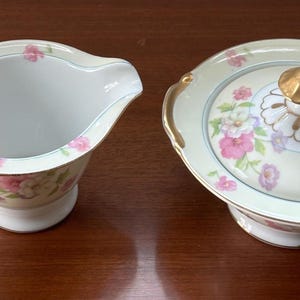 May include: A porcelain creamer and a lidded sugar bowl featuring a pale cream base with a delicate floral pattern of pink and purple flowers. Both pieces have gold-trimmed edges and handles, with the sugar bowl lid topped by a gold-colored knob.