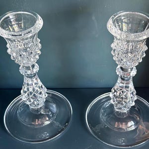May include: A pair of clear glass candle holders featuring a textured diamond pattern on the stems and bobeches. Each holder has a wide, smooth circular base and a flared top rim designed to support a taper candle in an upright position.