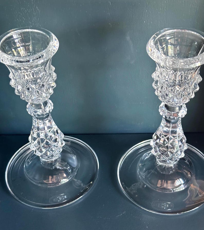 May include: A pair of clear glass candle holders featuring a textured diamond pattern on the stems and bobeches. Each holder has a wide, smooth circular base and a flared top rim designed to support a taper candle in an upright position.