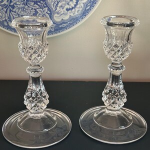 May include: A pair of clear glass candle holders featuring a faceted diamond-cut pattern on the upper cup and the central stem. Each holder has a wide, smooth, circular base and a flared top rim, crafted from transparent pressed glass material.