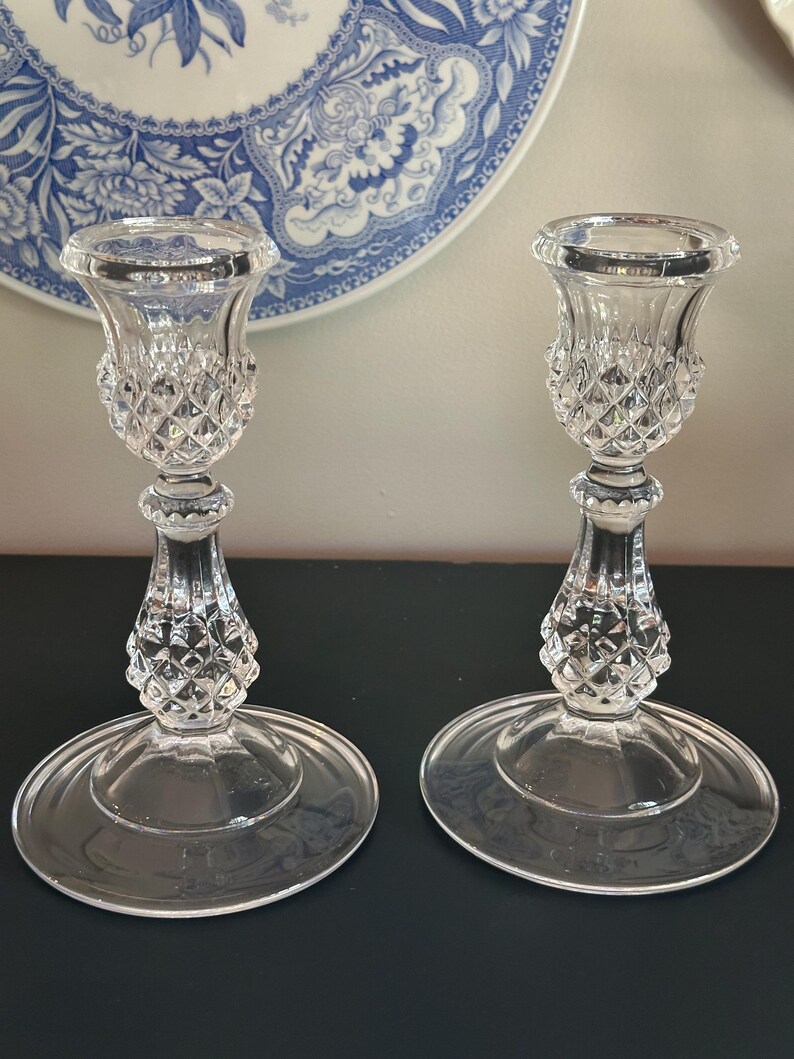 May include: A pair of clear glass candle holders featuring a faceted diamond-cut pattern on the upper cup and the central stem. Each holder has a wide, smooth, circular base and a flared top rim, crafted from transparent pressed glass material.