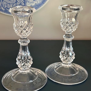 May include: A pair of clear glass candle holders featuring a faceted diamond-cut pattern on the upper cup and the central stem. Each holder has a wide, smooth circular base and a flared top rim designed to securely hold a standard taper candle.