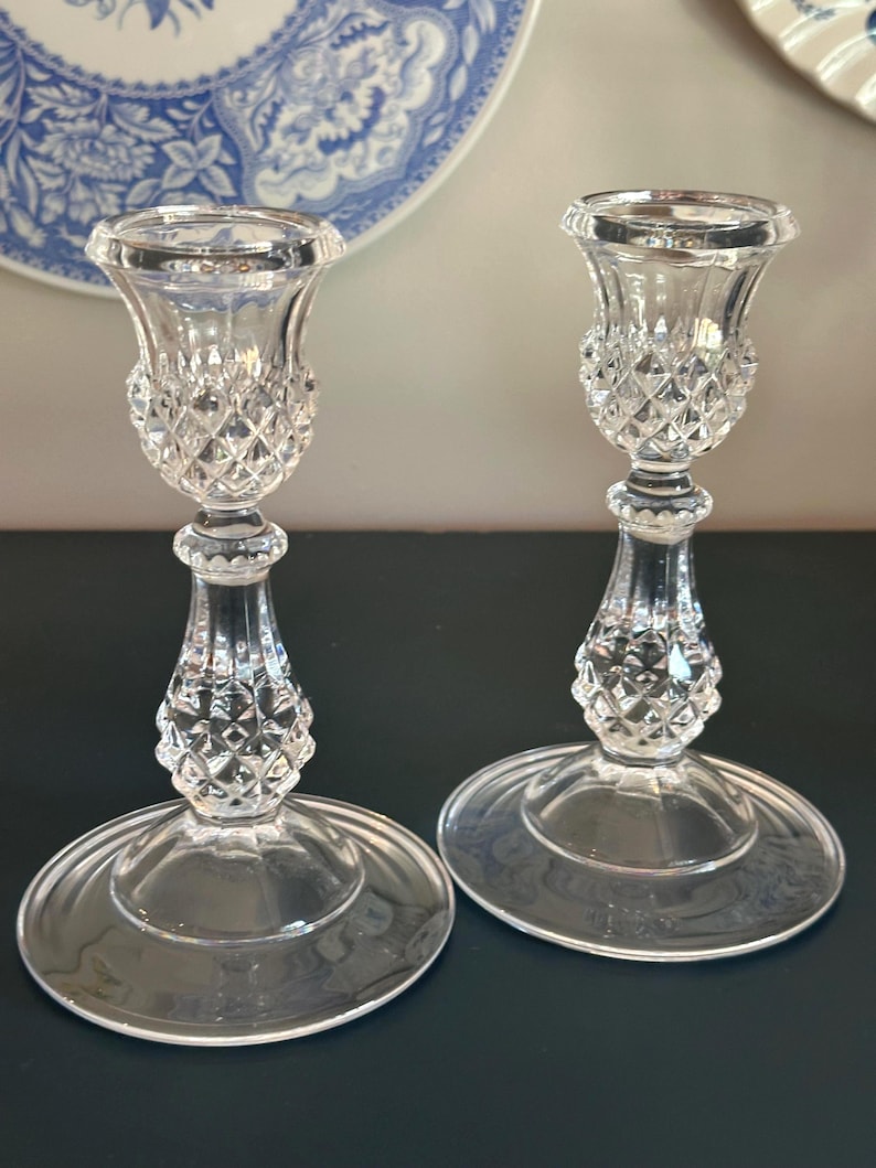 May include: A pair of clear glass candle holders featuring a faceted diamond-cut pattern on the upper cup and the central stem. Each holder has a wide, smooth circular base and a flared top rim designed to securely hold a standard taper candle.
