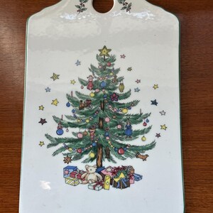 May include: White ceramic Christmas-themed cutting board with a green border and a hole for hanging. The board features a decorated Christmas tree with ornaments, stars, and presents. A teddy bear sits at the base of the tree.