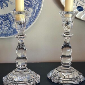 May include: A pair of clear glass candlesticks with a faceted, multi-tiered base and a central stem featuring rounded, bulbous sections. Each candlestick holds a single cream-coloured taper candle within a flared, textured top rim.
