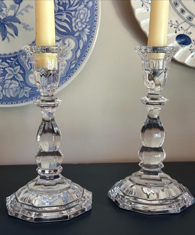 May include: A pair of clear glass candlesticks with a faceted, multi-tiered base and a central stem featuring rounded, bulbous sections. Each candlestick holds a single cream-coloured taper candle within a flared, textured top rim.