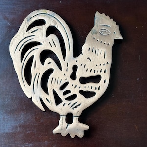 May include: A brass rooster-shaped trivet with a cutout design. The trivet features a detailed depiction of a rooster, with a textured surface and intricate cutouts. The color is a warm, antique brass.