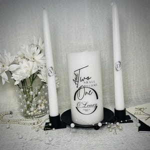 May include: Two white pillar candles with black calligraphy that reads "And Two Shall Become One" with the couple's last name and wedding date. The candles are on a black metal base.