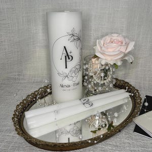 May include: Two white pillar candles with floral designs and the initials "A + P" and the text "Alexis - Paul 08.10.2024" on the larger candle. The candles are on a gold oval tray with a mirrored surface and white pearl beads.