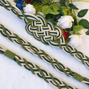 May include: A braided belt made of green and cream cotton rope with a knotted buckle. The belt is laid out on a white surface.