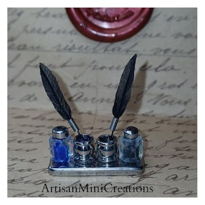 May include: Miniature inkwell set with two black feather quills. The set includes three inkwells and a clear glass bottle with blue ink. The base is silver-toned, and the background features handwritten script. The text "ArtisanMiniCreations" is visible.