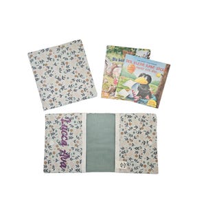 Könnte beinhalten: Ein Stoffbuchumschlag mit Blumenmuster in gedeckten Beige-, Blau- und Orangetönen, auf dem der Name Lucca Ava in violettem Garn gestickt ist. Das Set enthält eine passende Stoffbuchhülle und ein Kinderbuch mit dem Titel Der kleine Rabe Socke.