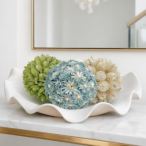 May include: A white, ruffled ceramic bowl holds three decorative spheres. The spheres are green, blue, and cream-colored, resembling floral arrangements. The bowl sits on a marble surface with a gold trim.