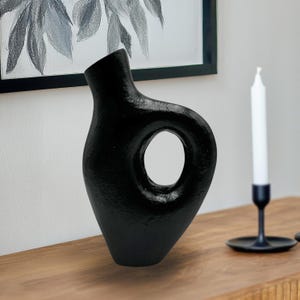 May include: Black ceramic vase with a unique, abstract design featuring a large, circular cutout. The vase has a textured, matte finish.