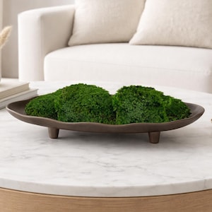 May include: A brown, oval-shaped decorative tray with three short legs, filled with vibrant green moss. The tray is placed on a white marble coffee table, creating a natural and elegant centerpiece.