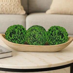 May include: Three green decorative moss balls sit in a light brown wooden bowl. The balls have a textured, spherical shape. The bowl is oval and rests on a white marble table. The background includes a beige pillow and a gray sofa.
