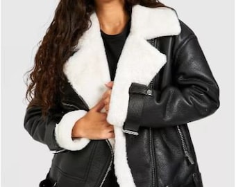 May include: A black jacket with a white faux shearling lining, featuring a wide notched collar, silver-toned metal zippers on the front and cuffs, and a buckled strap detail at the hem. The jacket has a textured finish and long sleeves.