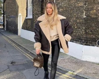 May include: A dark brown leather jacket with a thick beige shearling lining and collar, worn over a beige knit sweater and black leather-look leggings. The outfit is paired with black platform ankle boots and a small brown patterned backpack.