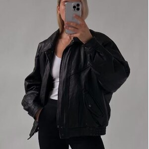 May include: A black leather bomber jacket with a zipper closure and a ribbed hem. The jacket has a relaxed fit and a slightly oversized silhouette.