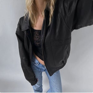 May include: A black leather jacket with a zipper closure, a collar, and pockets. The jacket is worn over a black lace top and blue jeans. The jacket has a bomber style with a ribbed hem and cuffs.