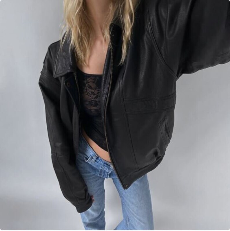 May include: A black leather jacket with a zipper closure, a collar, and pockets. The jacket is worn over a black lace top and blue jeans. The jacket has a bomber style with a ribbed hem and cuffs.