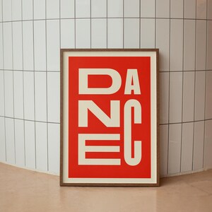 May include: A framed art print with the word "DANCE" in large, bold, off-white letters against a red background. The artwork is displayed against a white tiled wall and a light brown floor.