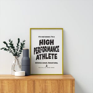 May include: A framed print with a yellow frame and a white background. The text on the print reads "It's real sweat. I'm a HIGH PERFORMANCE ATHLETE. Athletes sweat. Sweat baby. Ki ki ki, ra, sweat sweat."