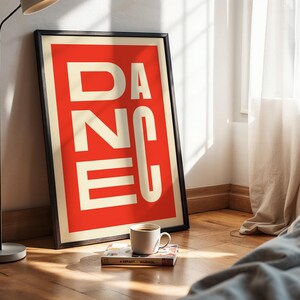 May include: A framed art print with the word "DANCE" in white block letters against a red background. The print is propped against a wall on a wooden floor, with a coffee cup and a book nearby.