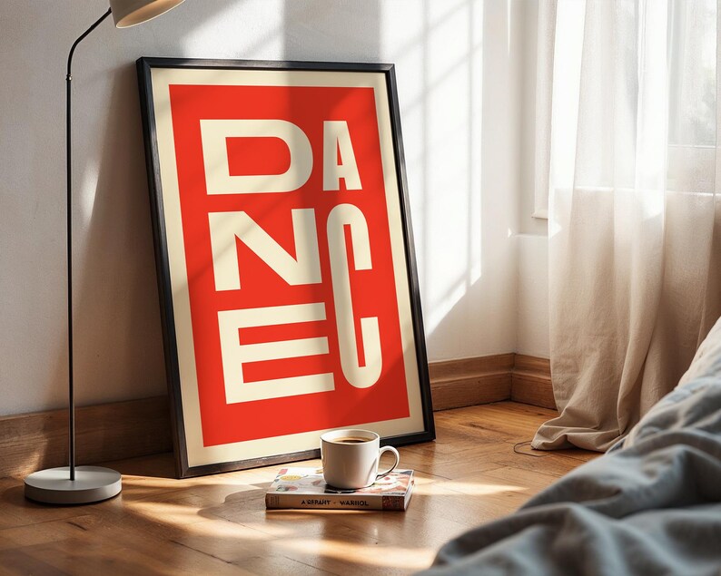 May include: A framed art print with the word "DANCE" in white block letters against a red background. The print is propped against a wall on a wooden floor, with a coffee cup and a book nearby.