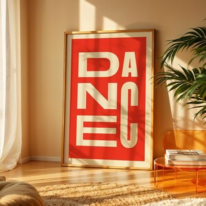 May include: A framed art print with the word "DANCE" in large, white, block letters against a red background. The print is in a gold frame and leans against a wall. A potted plant and a small table with books are in the scene.