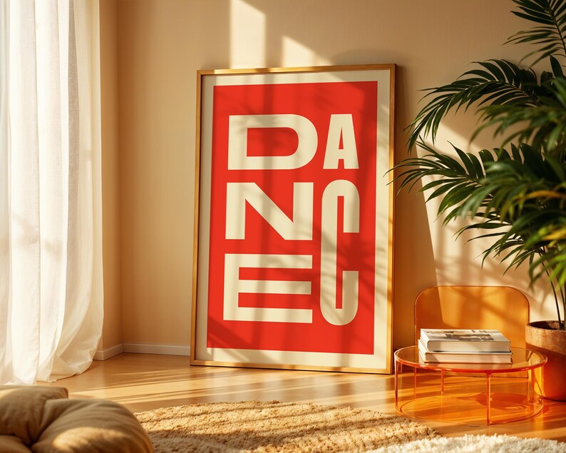 May include: A framed art print with the word "DANCE" in large, white, block letters against a red background. The print is in a gold frame and leans against a wall. A potted plant and a small table with books are in the scene.